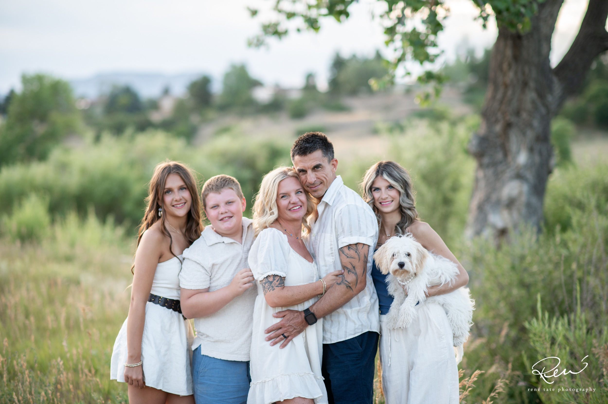 a family of five stand together in an open field of golden grass with their family dog and mountains in the background in Colorado Springs CO