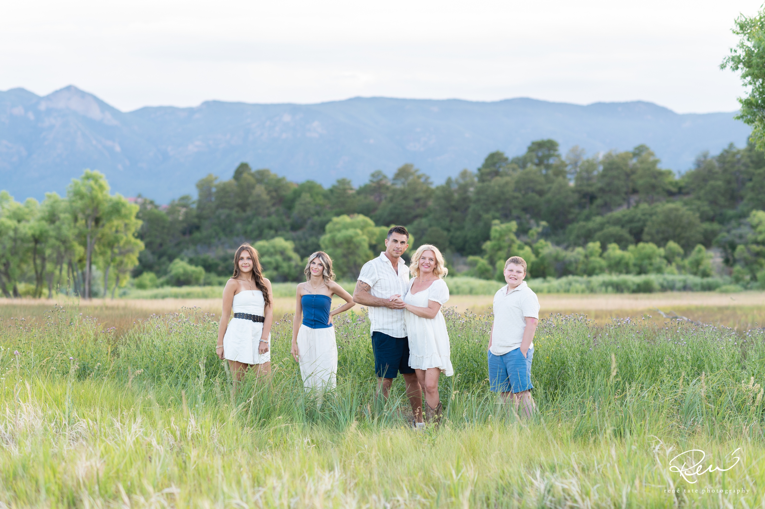 a family of five stand together in an open field of golden grass with their family dog and mountains in the background in Colorado Springs CO