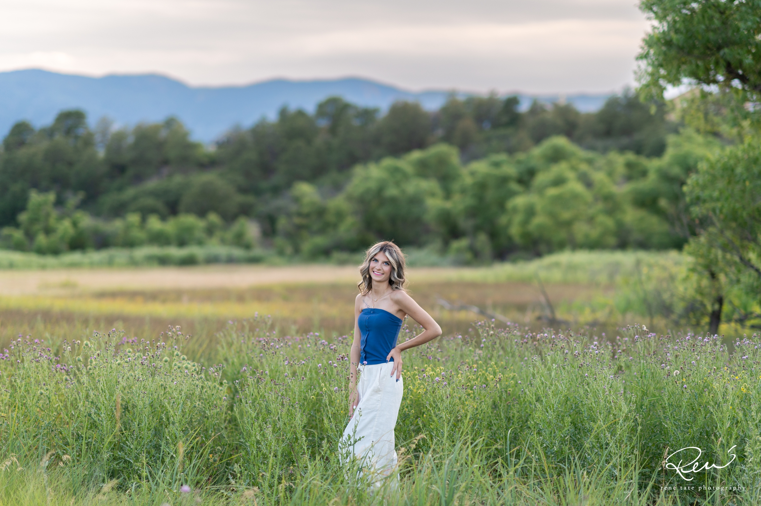 High school senior girl poses for the camera in an open field of flowers with mountains in the background in Colorado Springsds 