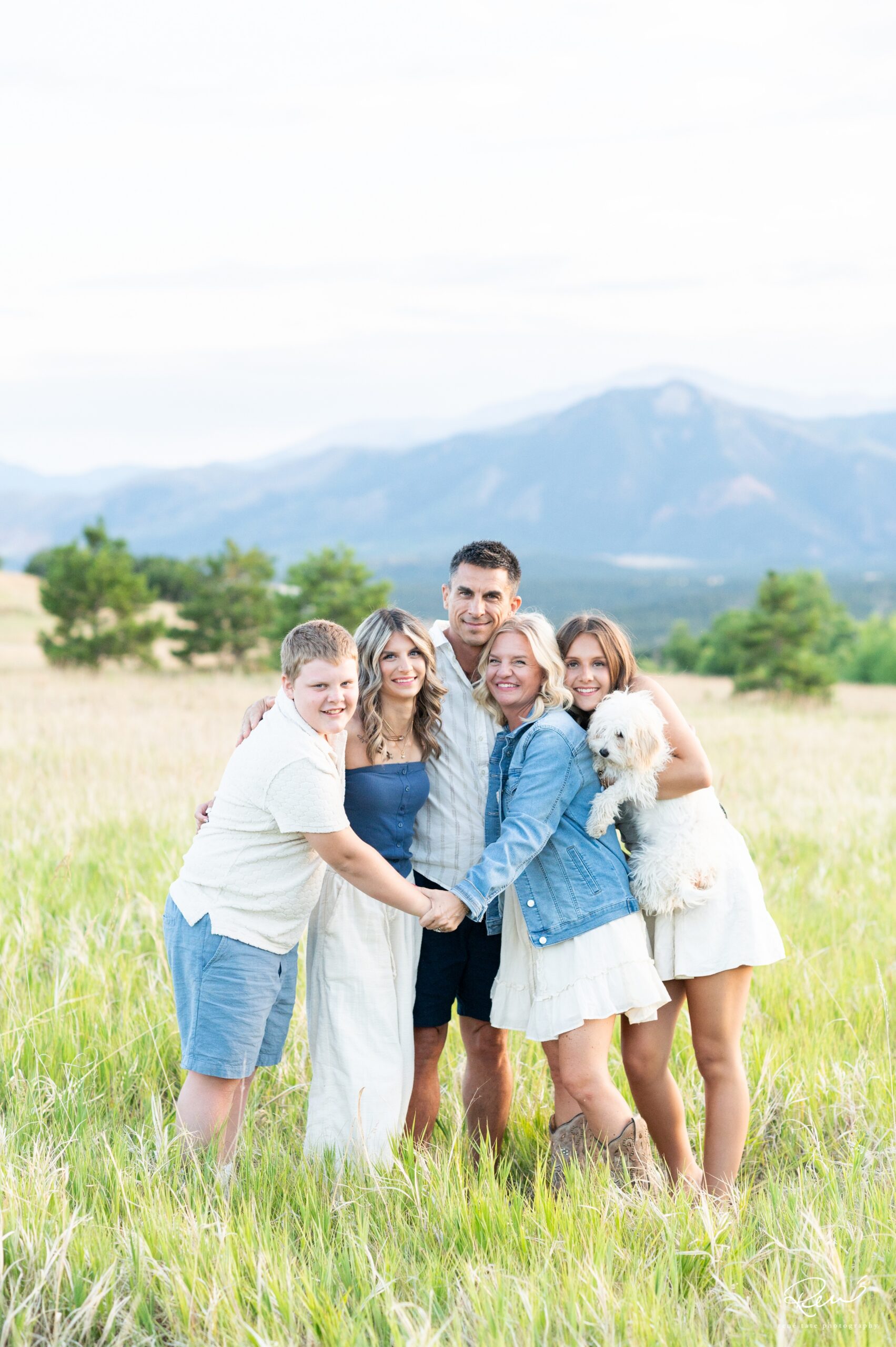 Family of five with two teenage daughters and a preteen son and their family dog, standing in an open field with a mountain background in Colorado Springs at sunset 
